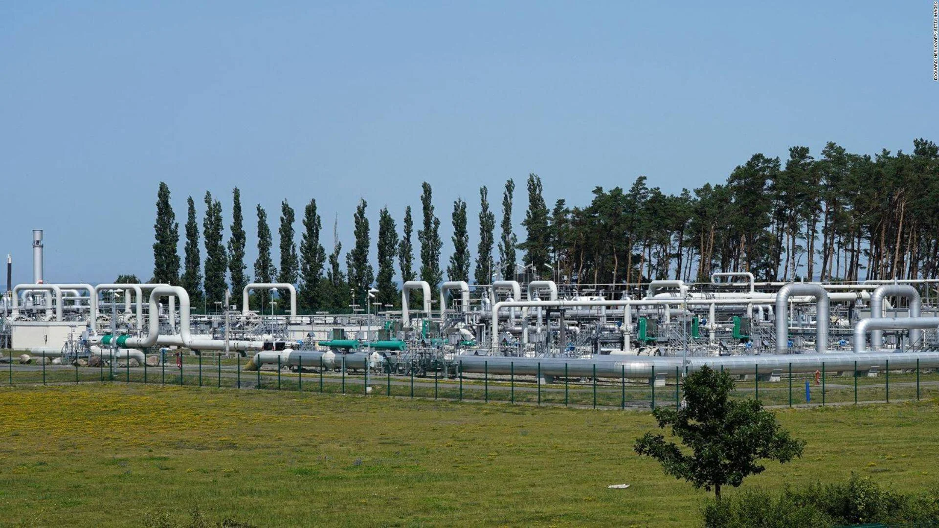 Estación de recepción de gasoducto con extensas tuberías metálicas grises y verdes rodeada de árboles y pradera.