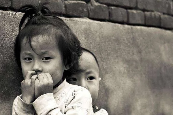 Dos niños pequeños de aspecto asiático junto a una pared de ladrillos, en fotografía blanco y negro, con expresión tímida.