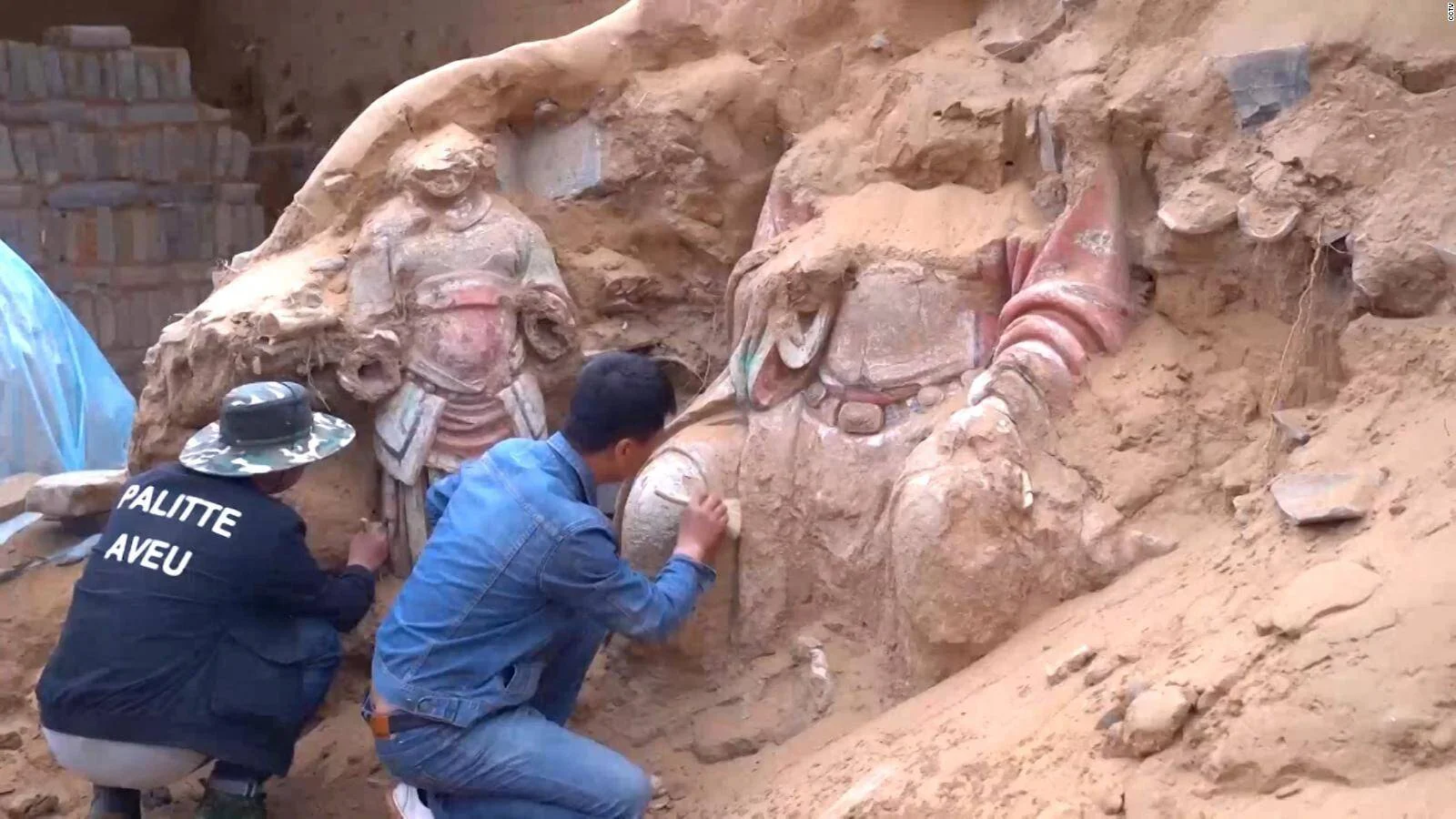 Dos arqueólogos examinan figuras de terracota pintadas con colores parcialmente enterradas en sedimento durante una excavación.