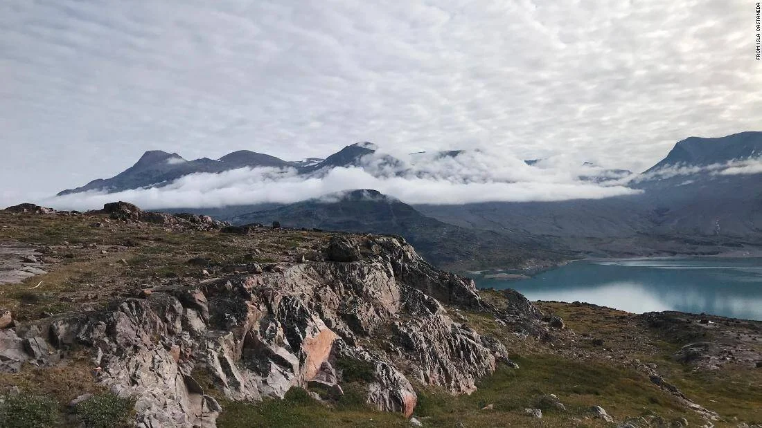 Vista panorámica de montañas cubiertas de nubes bajas junto a un fiordo de aguas turquesas en un paisaje ártico rocoso.