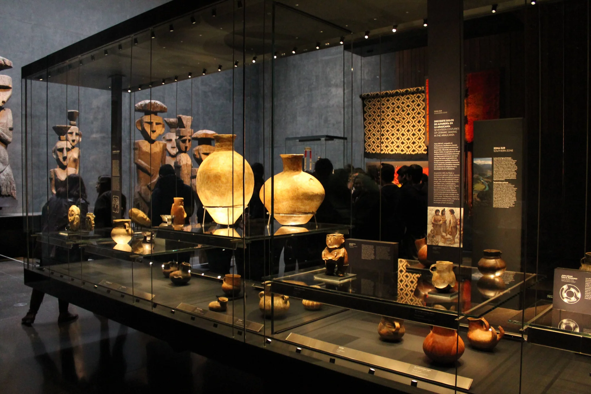 Vitrina de museo con cerámica precolombina y esculturas de madera mapuche iluminadas, con visitantes al fondo.