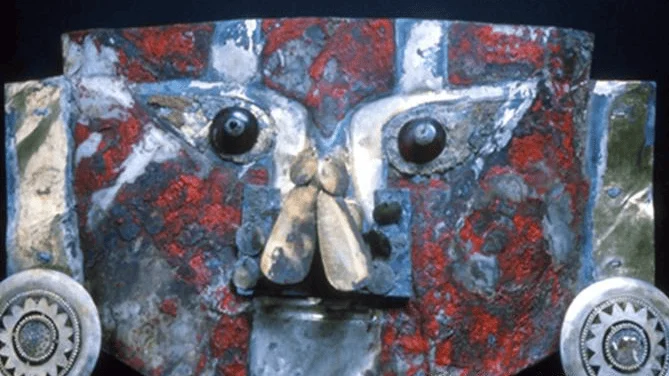 Máscara ceremonial prehispánica de metal con restos de pigmento rojo, ojos esféricos oscuros y ornamentos circulares laterales.