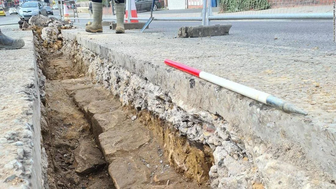 Zanja abierta en una acera urbana expone una antigua estructura de piedra enterrada bajo el pavimento moderno.