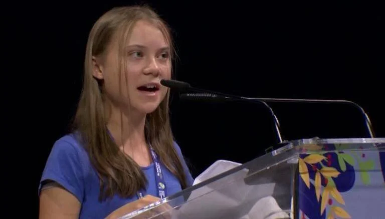 Una joven con remera azul habla en un podio transparente con micrófono ante un auditorio.