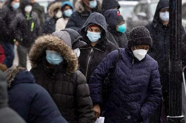 Grupo de personas con mascarillas haciendo fila bajo una nevada, vestidas con abrigos de invierno.