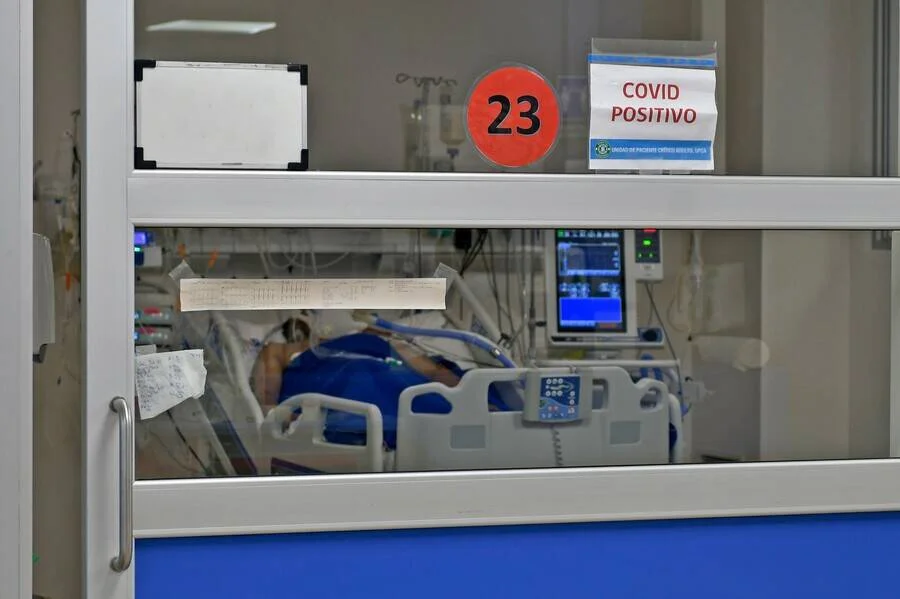 Paciente internado en cama hospitalaria visto tras la puerta de vidrio de una sala con cartel de Covid Positivo y número 23.