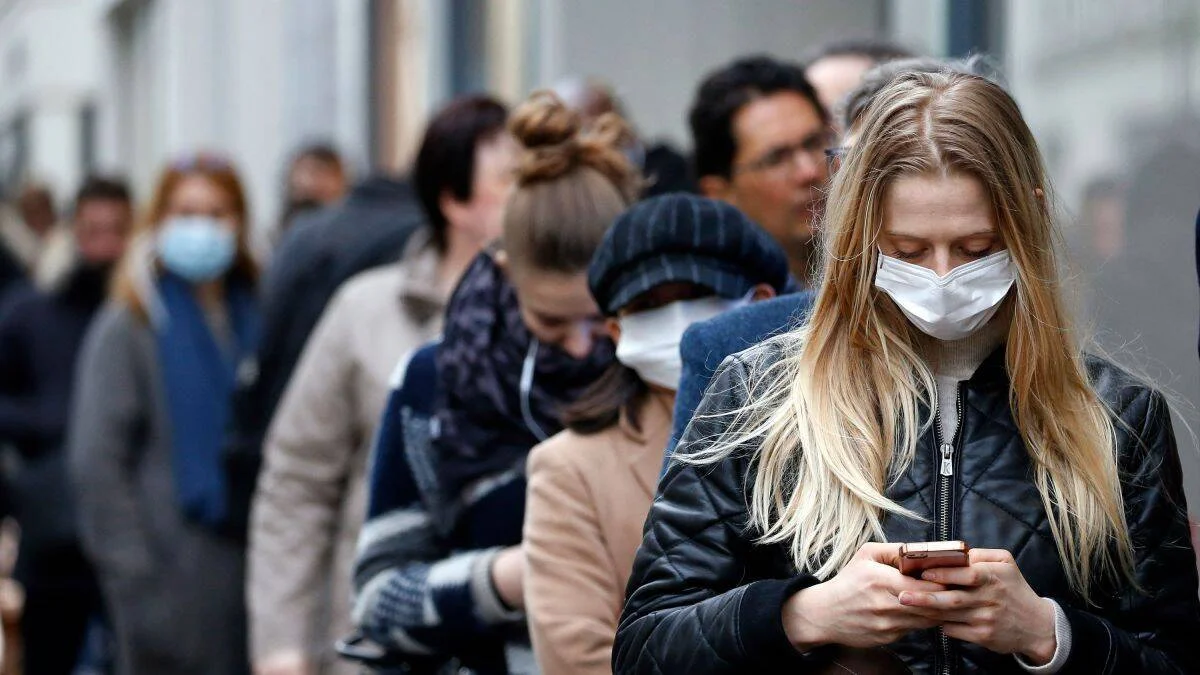 Grupo de personas con mascarillas sanitarias aguarda en fila en una calle urbana; una mujer al frente consulta su teléfono.