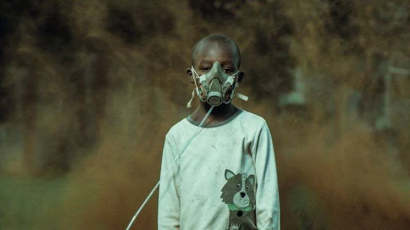 Niño con mascarilla respiratoria de pie en un entorno lleno de polvo o humo, evocando contaminación ambiental.