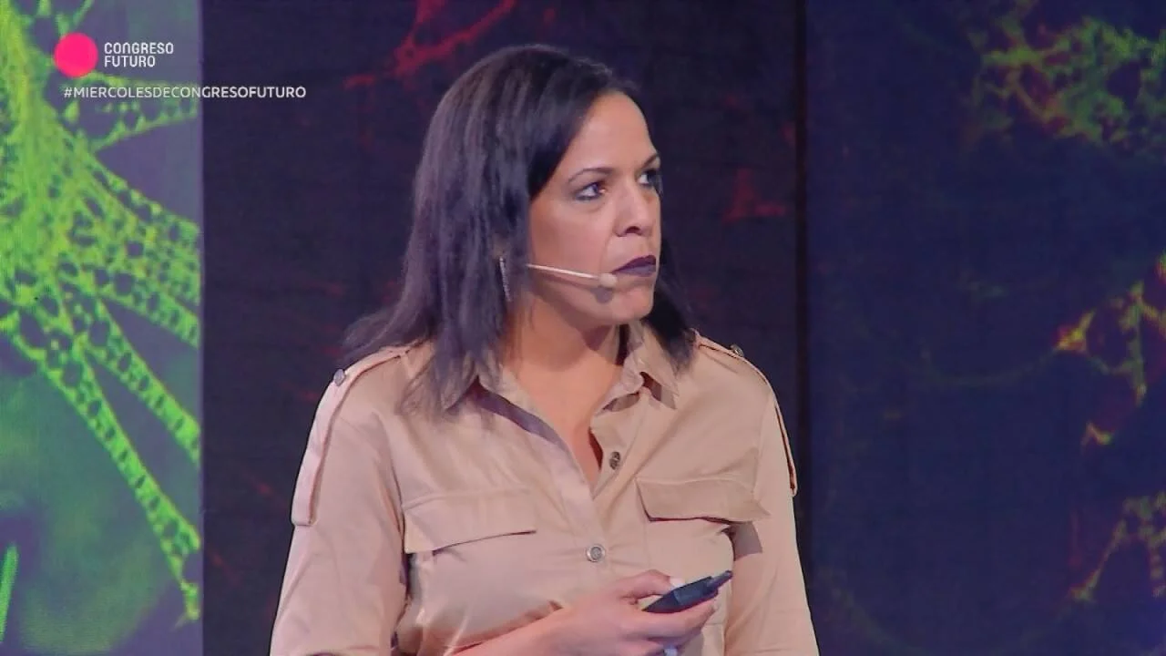 Mujer con micrófono de diadema expone en el escenario del evento Congreso Futuro sosteniendo un control remoto.