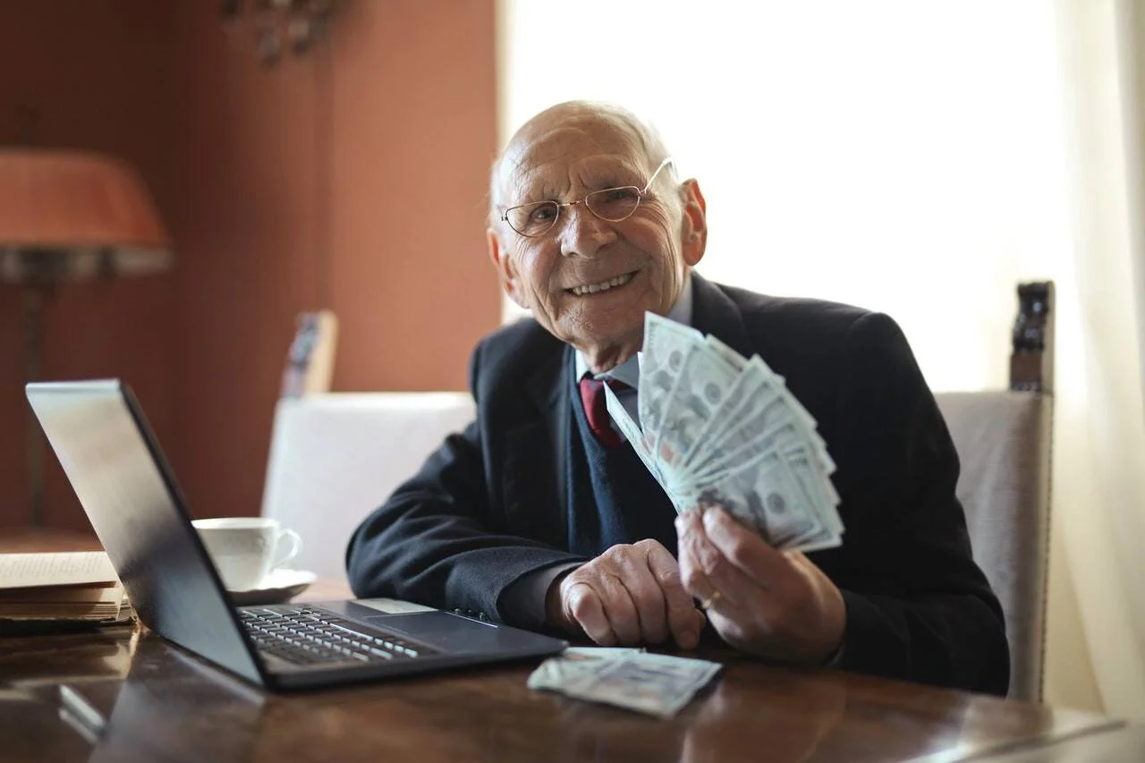 Hombre mayor con traje y gafas muestra un abanico de billetes sentado frente a una laptop en un escritorio.