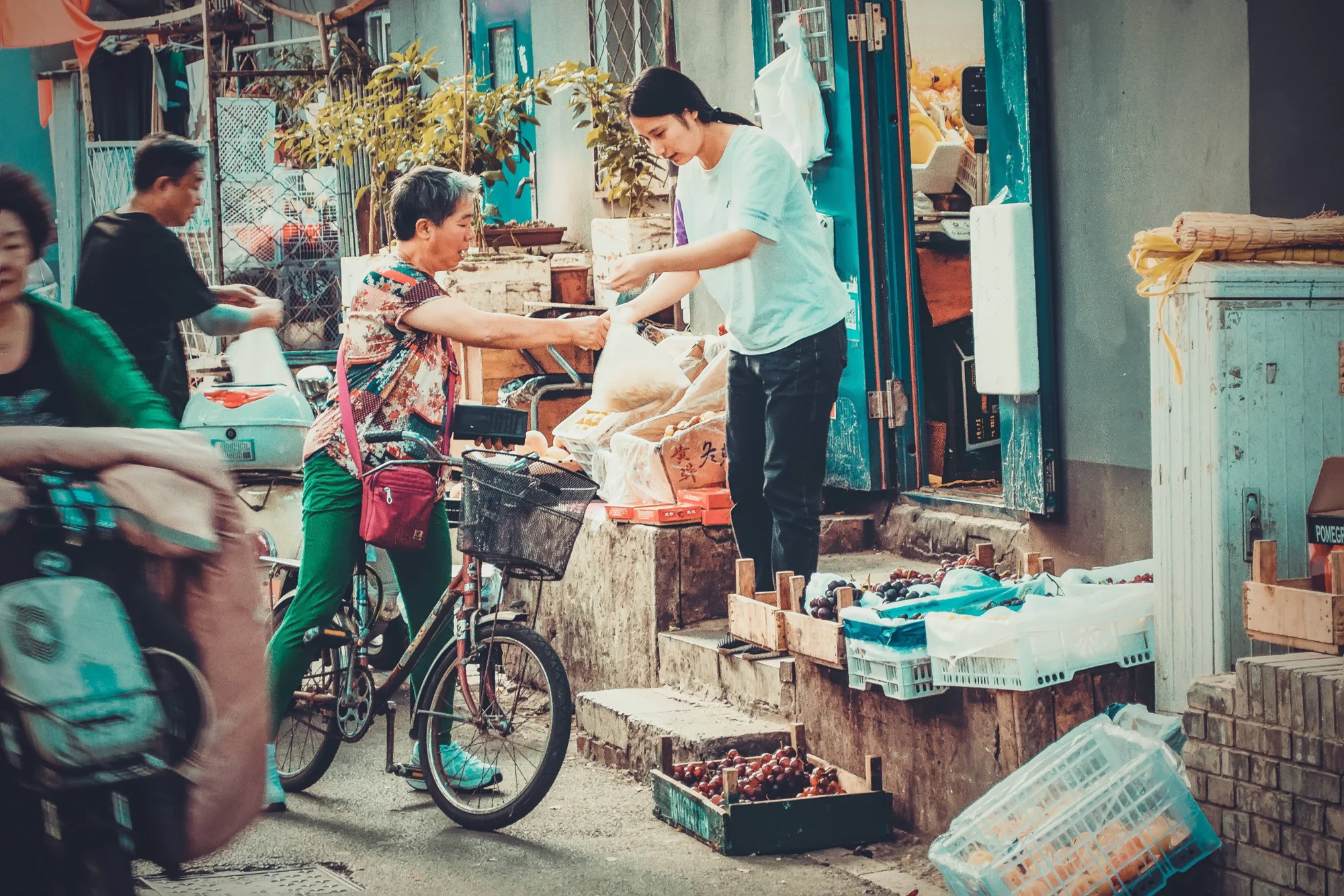 Una mujer mayor en bicicleta recibe un paquete de una vendedora en un puesto de frutas callejero en Asia.