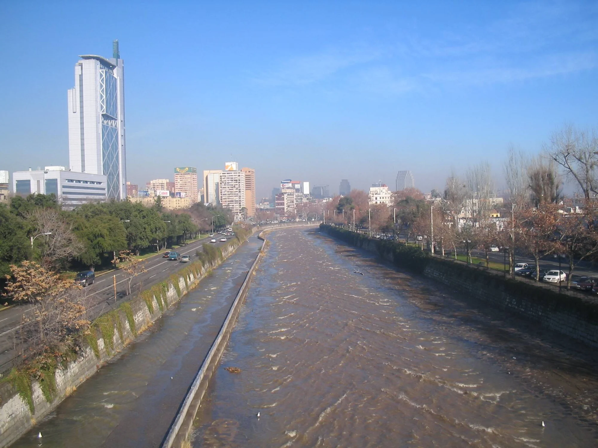 Río de aguas turbias y crecidas atraviesa una ciudad con edificios altos y avenidas flanqueadas por árboles.