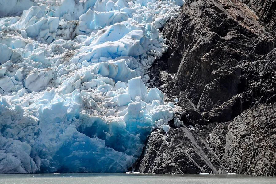 Frente de un glaciar con hielo azul fragmentado en contacto con una escarpada pared de roca oscura sobre el agua.