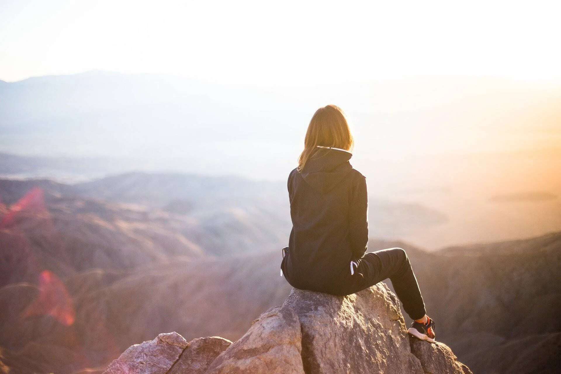 Una mujer de espaldas sentada sobre una roca en la cima de una montaña, contemplando un vasto paisaje iluminado por la luz del atardecer.