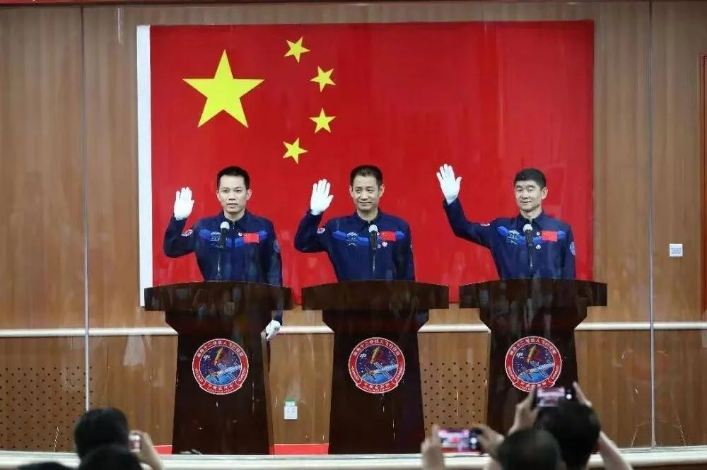 Tres taikonautas con trajes azules saludan desde sus atriles ante la bandera de China en una conferencia previa al lanzamiento.