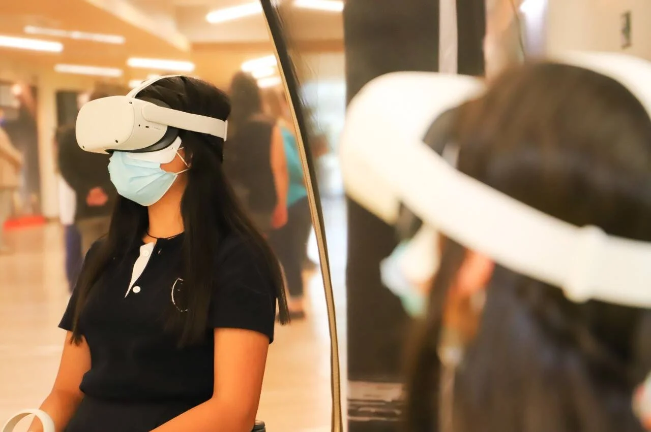 Mujer con mascarilla quirúrgica y visor de realidad virtual sentada en un pasillo hospitalario, con su reflejo visible en un espejo.
