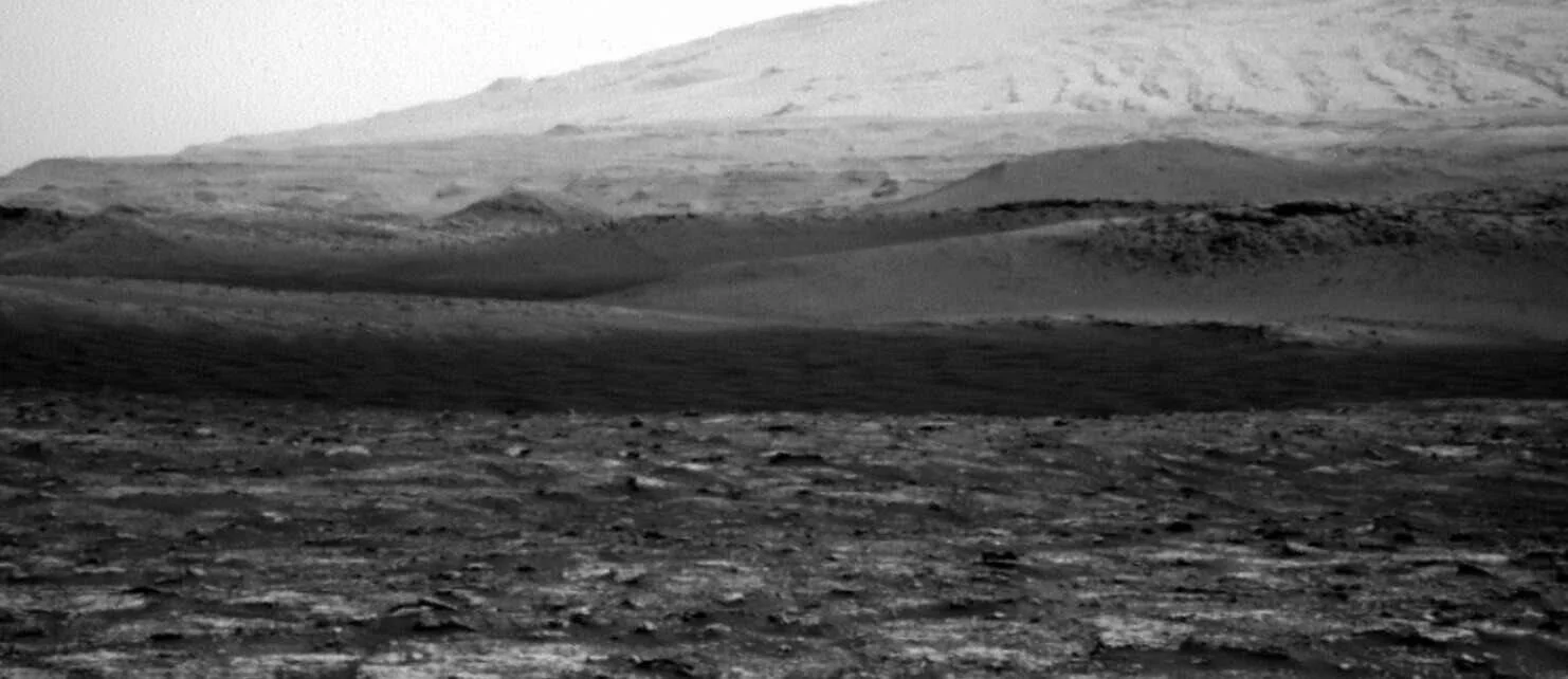 Fotografía en blanco y negro de la superficie marciana con terreno rocoso en primer plano, dunas y una gran montaña al fondo.