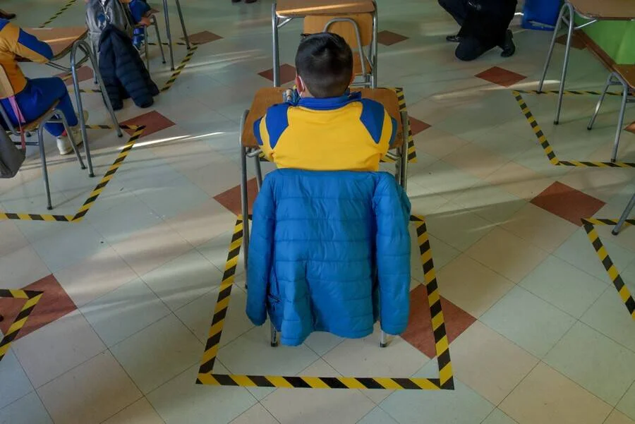Alumno sentado de espaldas en un aula con cuadrados de cinta señalizadora en el piso para garantizar el distanciamiento social.