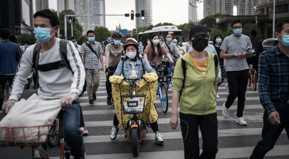 Decenas de personas con mascarillas sanitarias cruzan una concurrida intersección urbana en bicicleta y a pie.