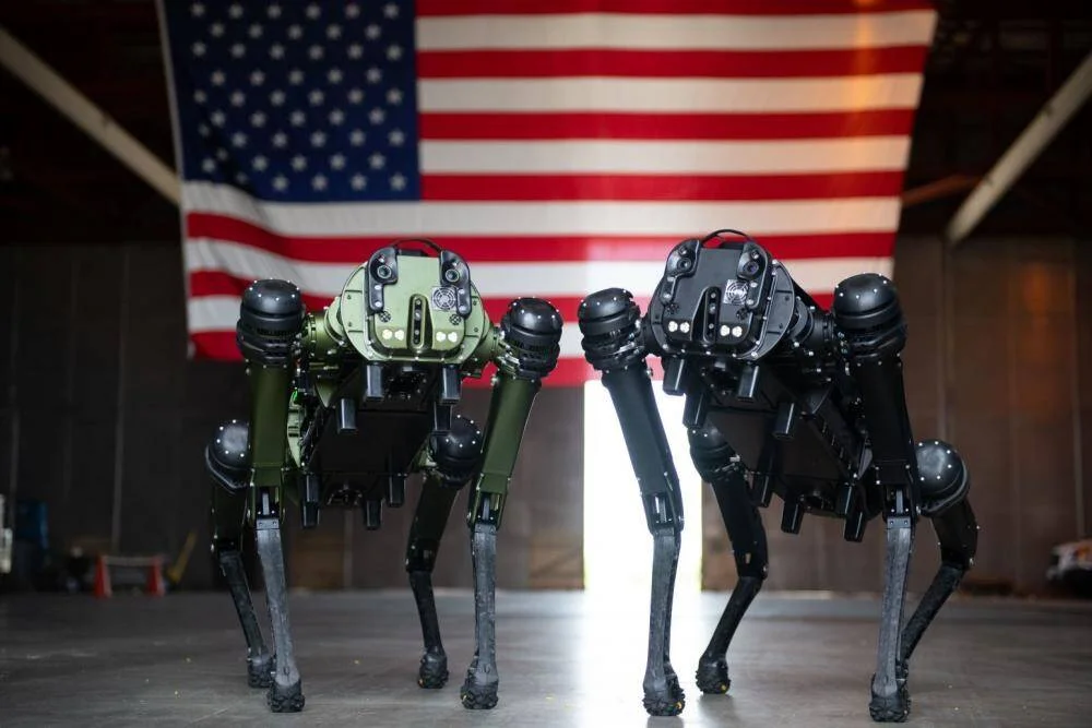 Dos robots cuadrúpedos de uso militar, uno verde oliva y otro negro, frente a una bandera de Estados Unidos.