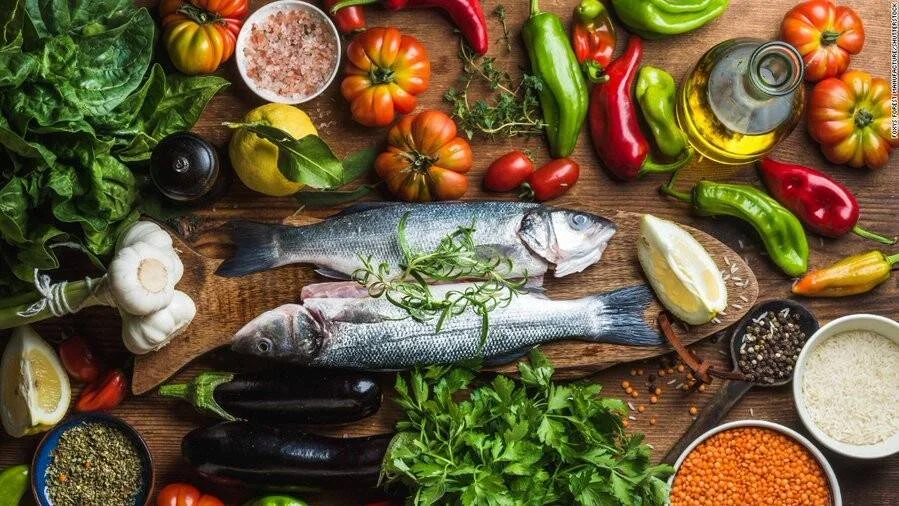 Dos pescados crudos sobre tabla de madera rodeados de verduras, hierbas, especias, aceite de oliva y legumbres frescos.