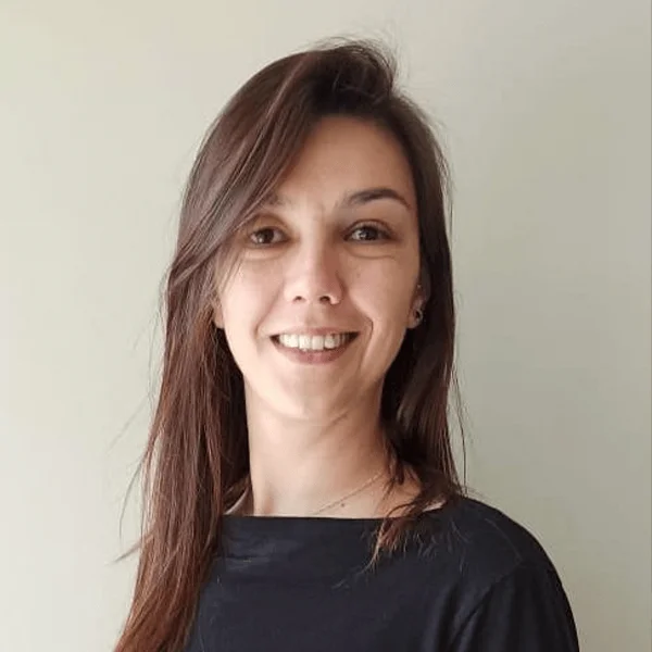 Mujer joven de cabello largo castaño sonriendo, vestida con remera negra, sobre fondo gris claro.
