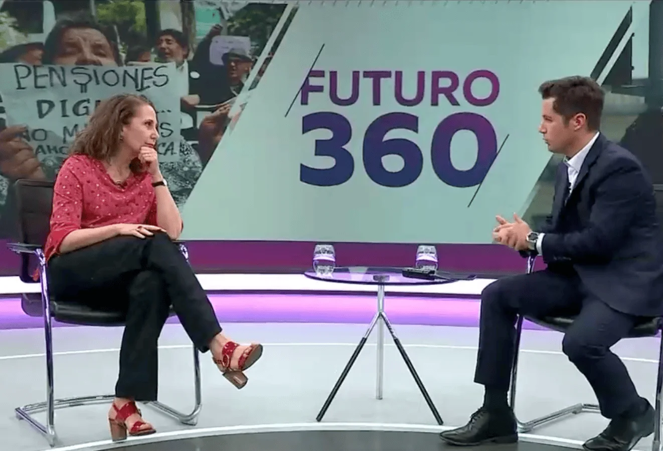 Una mujer y un hombre conversan en el estudio del programa televisivo Futuro 360, con imagen de protesta por pensiones dignas al fondo.