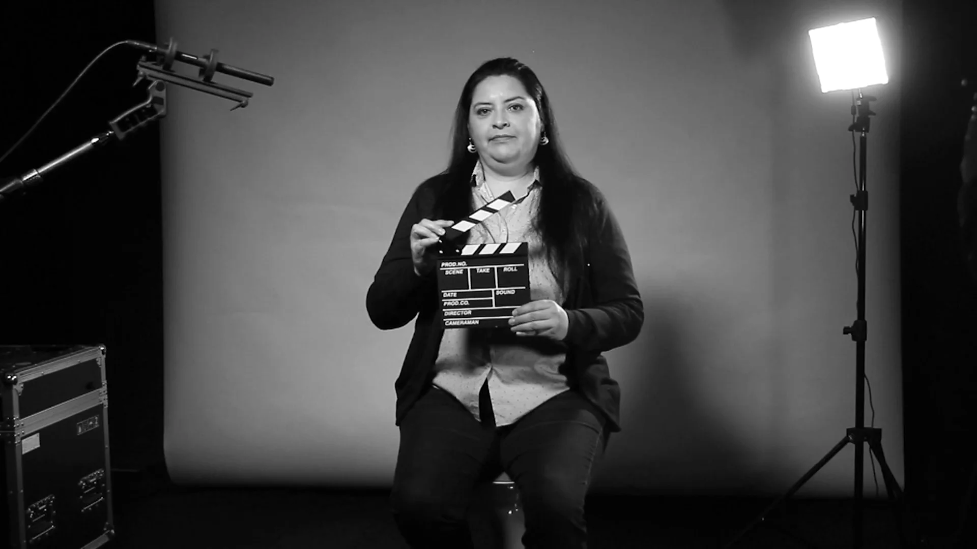 Mujer sentada en un estudio de filmación sostiene una claqueta de cine, rodeada de equipo de iluminación, en blanco y negro.