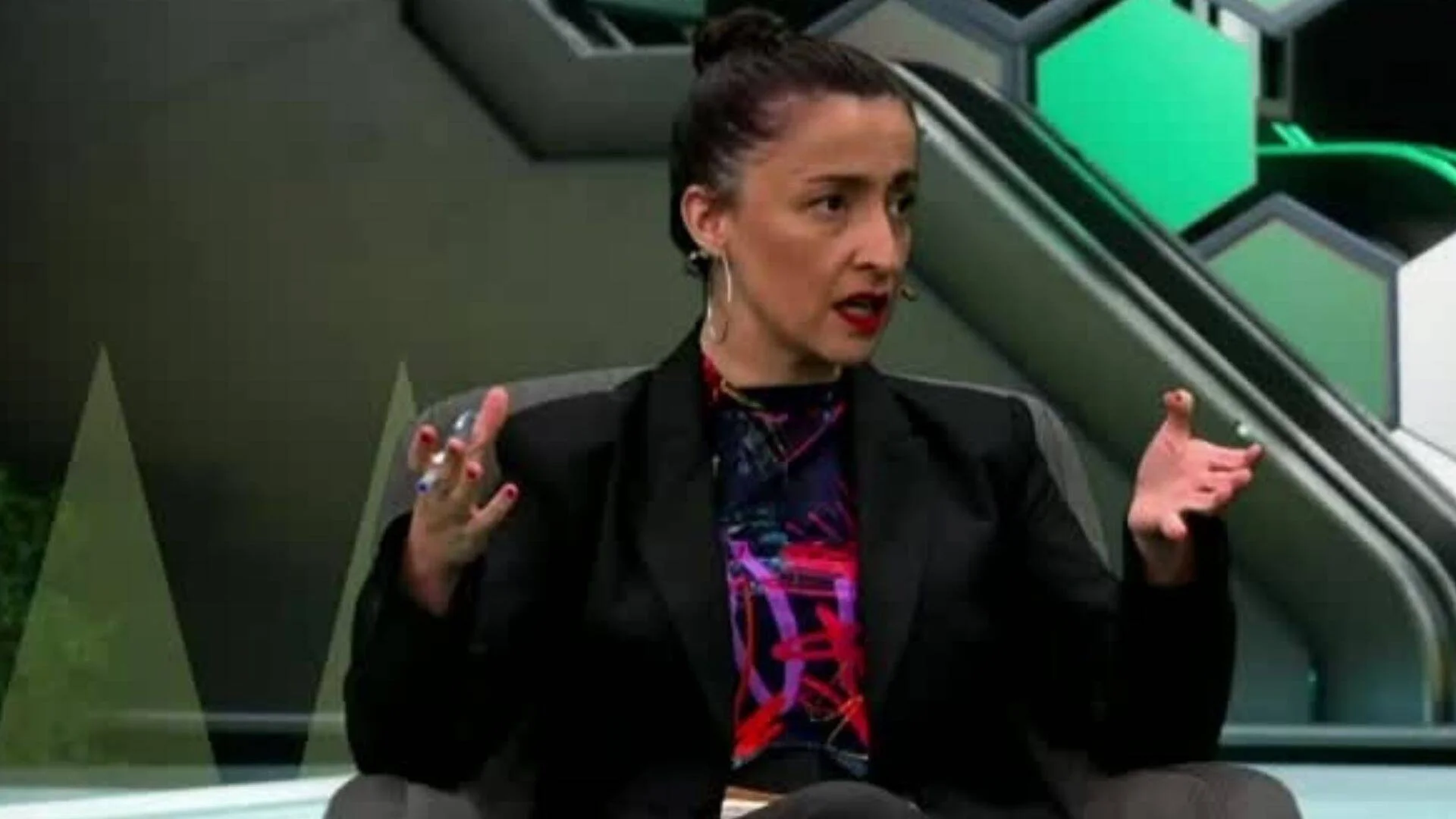 Mujer con blazer negro y blusa estampada gesticula mientras habla en un plató de televisión con decorado verde y gris.