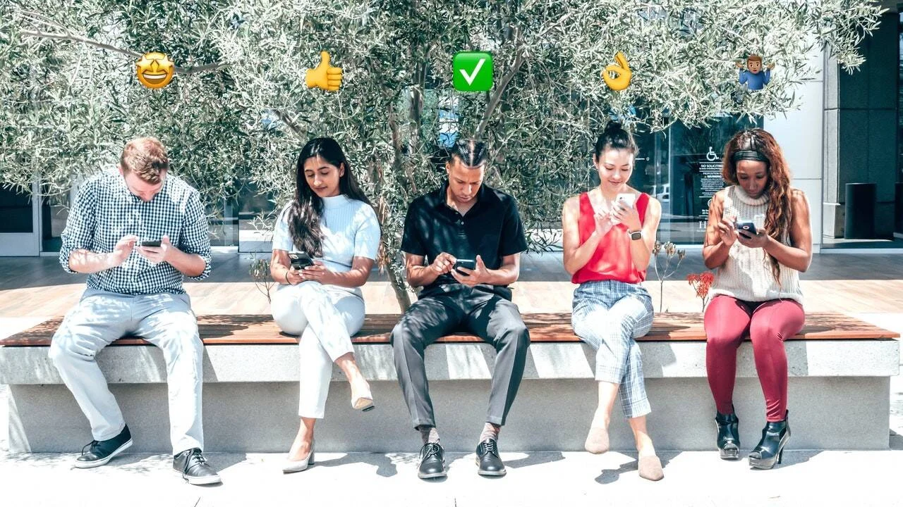 Cinco jóvenes sentados en un banco al aire libre mirando sus teléfonos móviles, con emojis positivos superpuestos sobre sus cabezas.