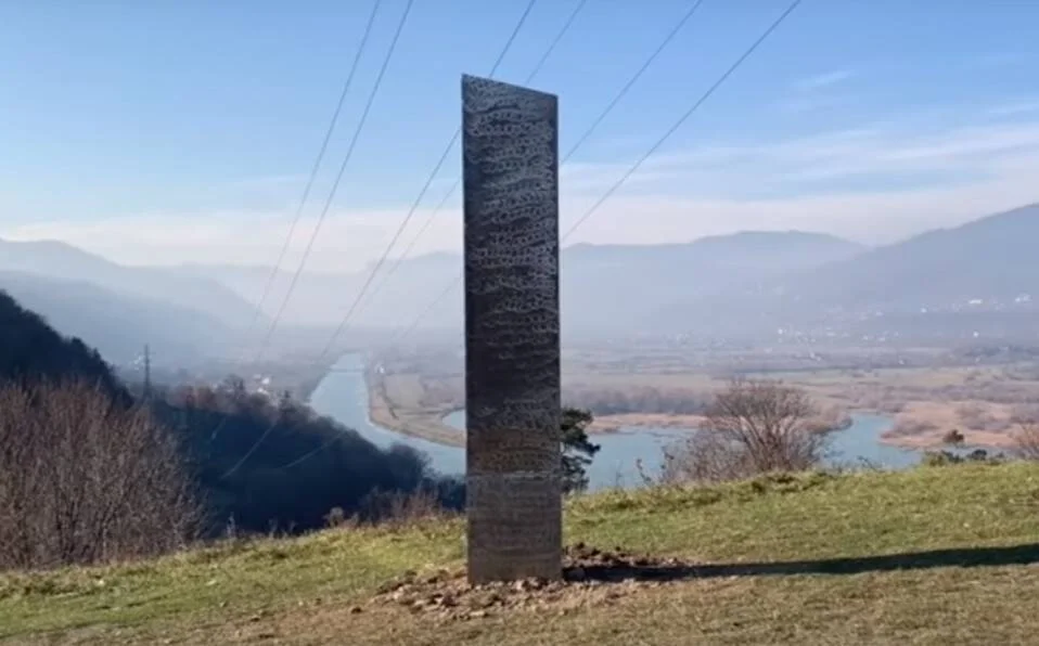 Monolito metálico oscuro erguido en una colina con un río y montañas de fondo bajo cielo azul.