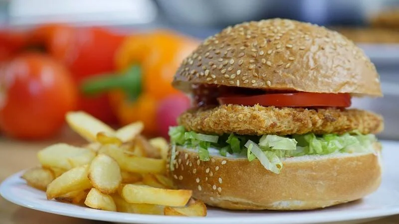 Hamburguesa de pollo empanado con lechuga y tomate en pan de sésamo, acompañada de papas fritas en plato blanco.