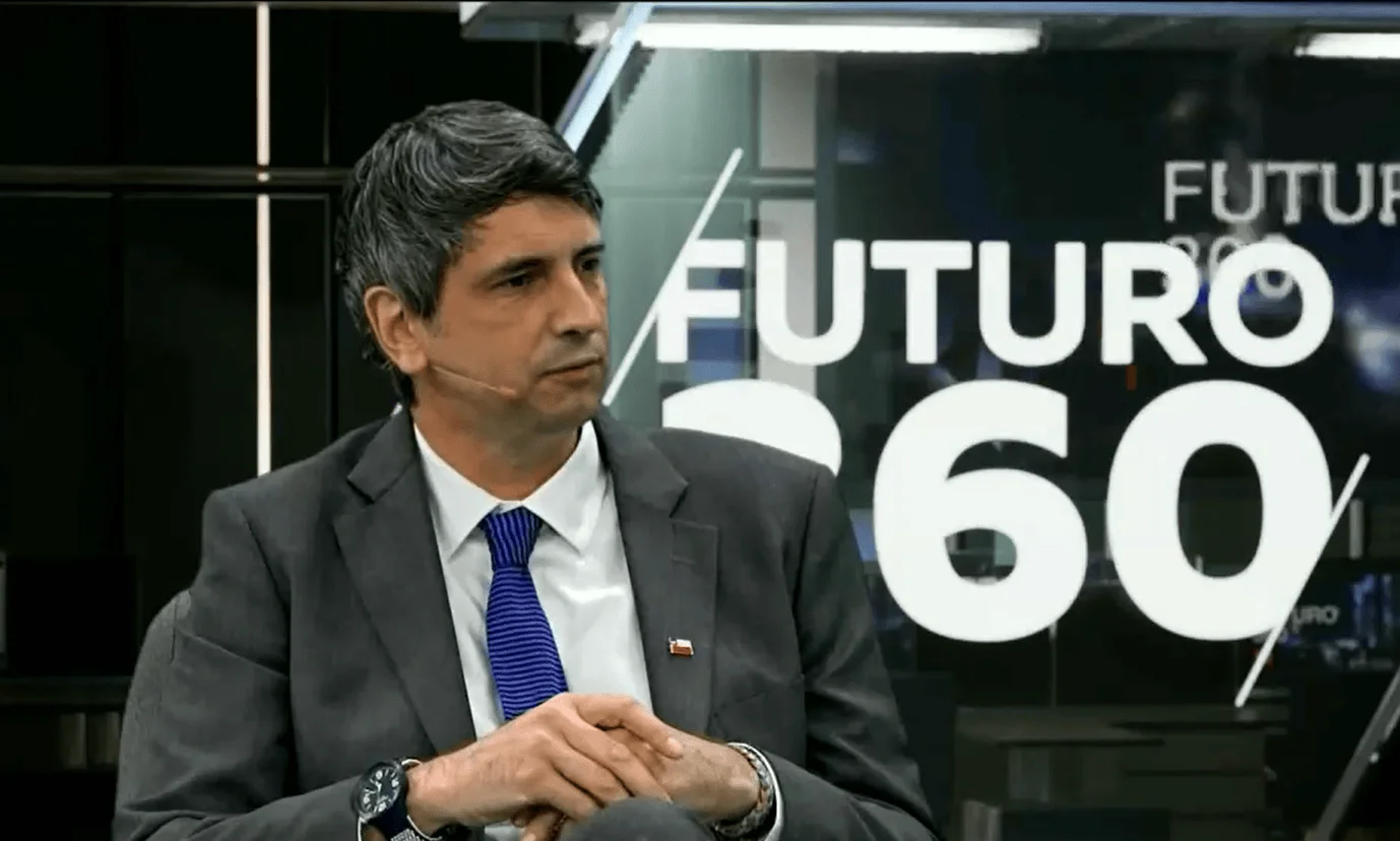 Hombre con traje gris y corbata azul, con pin de la bandera chilena, sentado durante una entrevista en el programa Futuro 360.