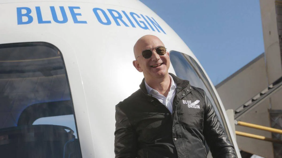 Hombre con gafas de sol y chaqueta de cuero de Blue Origin sonríe frente a una cápsula espacial de esa compañía.