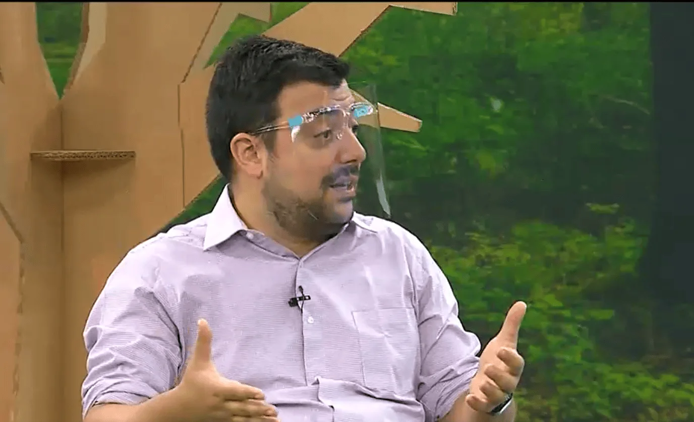 Hombre con anteojos y camisa clara gesticula mientras habla en un estudio de televisión con fondo verde y decorado de cartón.