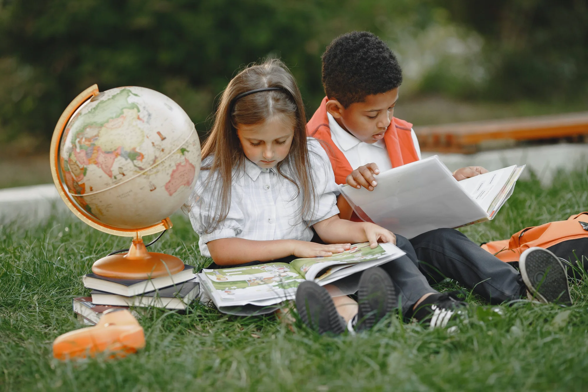 Dos niños en edad escolar leen libros sentados en el césped junto a un globo terráqueo y una pila de libros.