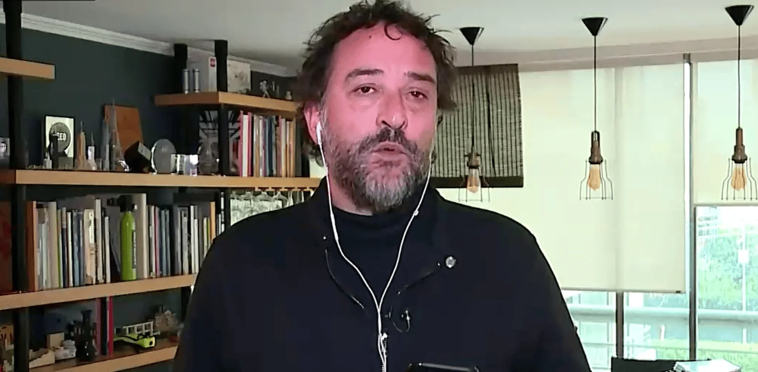 Hombre de mediana edad con barba y auriculares blancos habla frente a una biblioteca, durante una entrevista remota desde su hogar.
