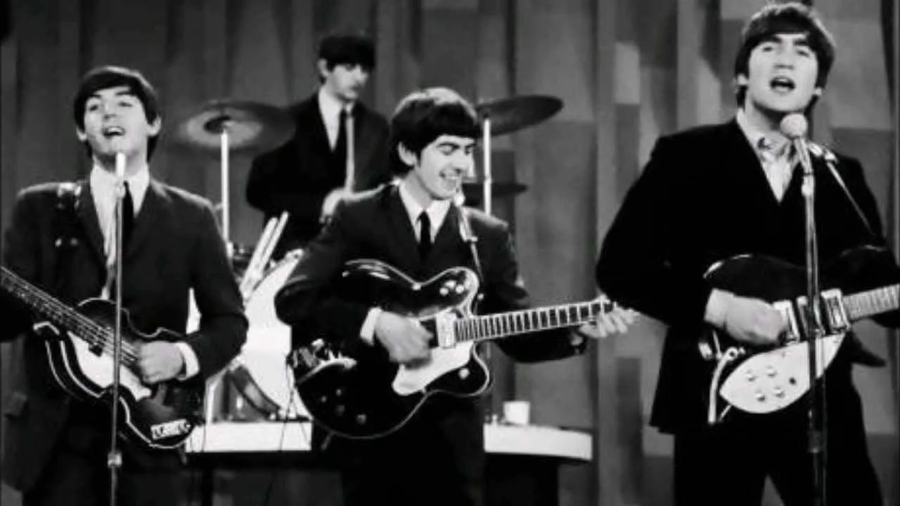 Cuatro jóvenes músicos con trajes oscuros tocan guitarras eléctricas y batería en un escenario televisivo, en blanco y negro.