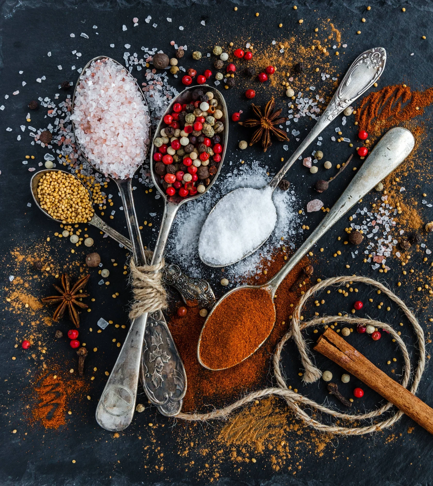Cucharas de plata antiguas con sal rosa del Himalaya, pimienta mixta, sal blanca y páprika, rodeadas de anís estrellado, canela y semillas de mostaza sobre fondo oscuro.