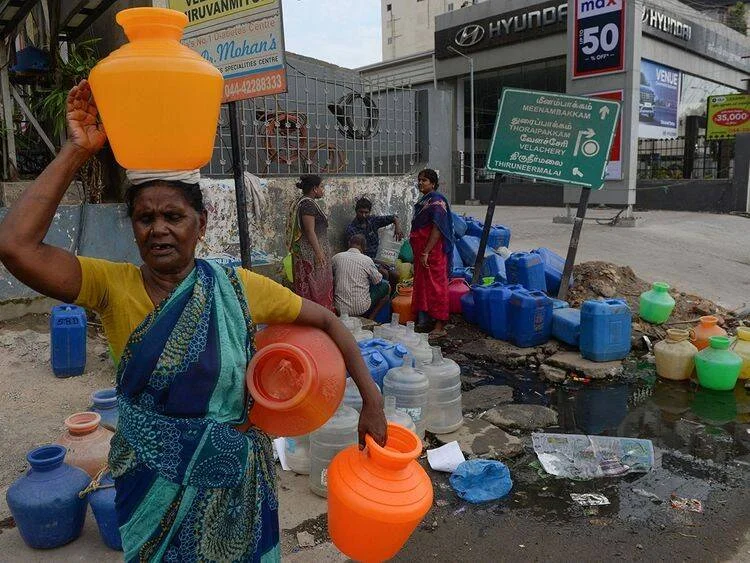 Una mujer carga recipientes plásticos con agua en una calle de Chennai, India, donde vecinos se abastecen ante la escasez hídrica.