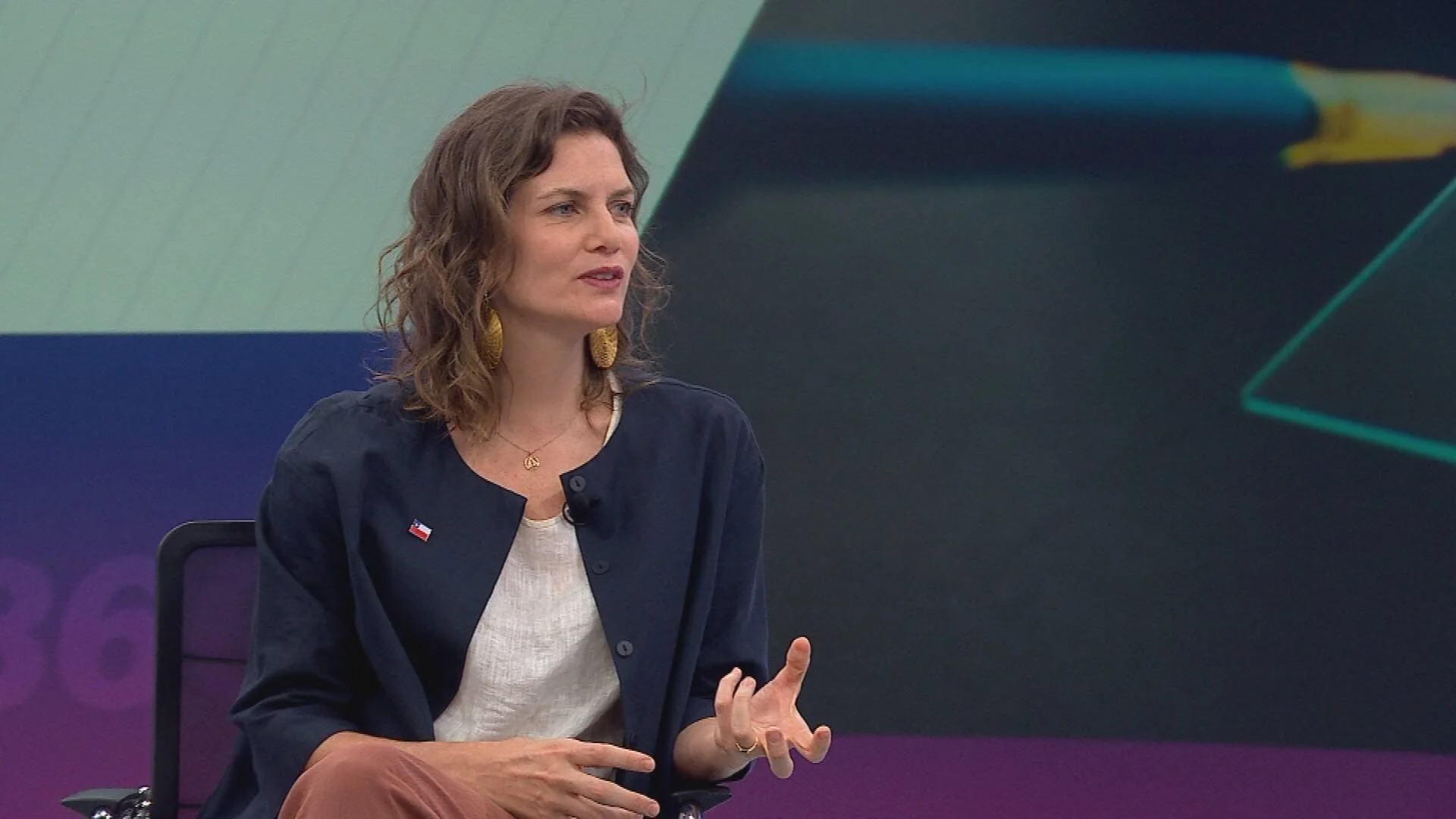 Mujer con chaqueta azul marino y pin de la bandera chilena habla gesticulando en un escenario de conferencia.
