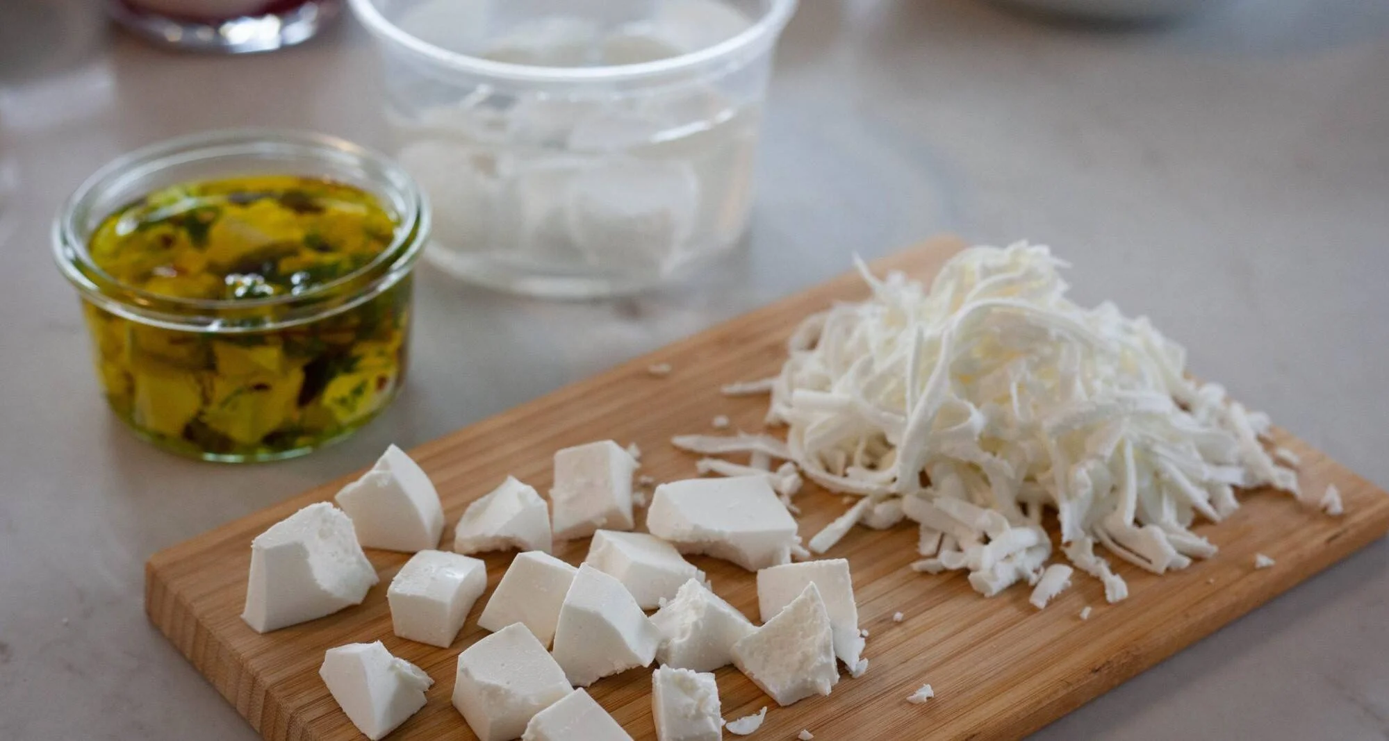 Tabla de madera con queso blanco cortado en cubos y rallado, junto a un frasco de aceite de oliva con hierbas.