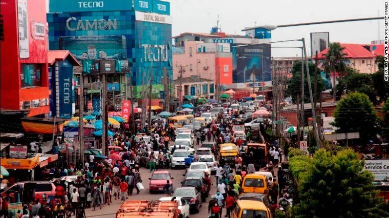 Avenida urbana abarrotada de personas y vehículos junto a edificios con grandes vallas publicitarias de la marca Tecno.