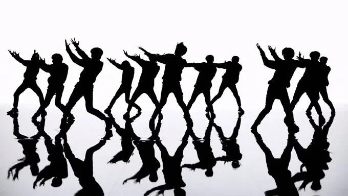 Siluetas en negro de un grupo numeroso de personas bailando sincronizadas, reflejadas en un suelo brillante sobre fondo blanco.
