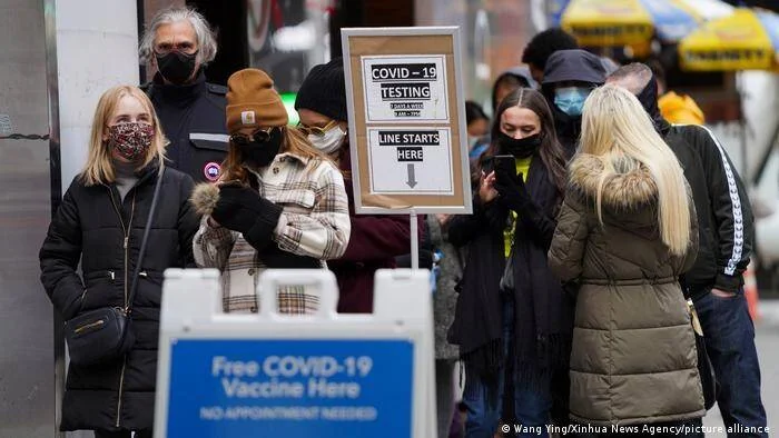 Personas con mascarilla hacen fila junto a carteles de testeo y vacunación gratuita contra el COVID-19 en una calle urbana.