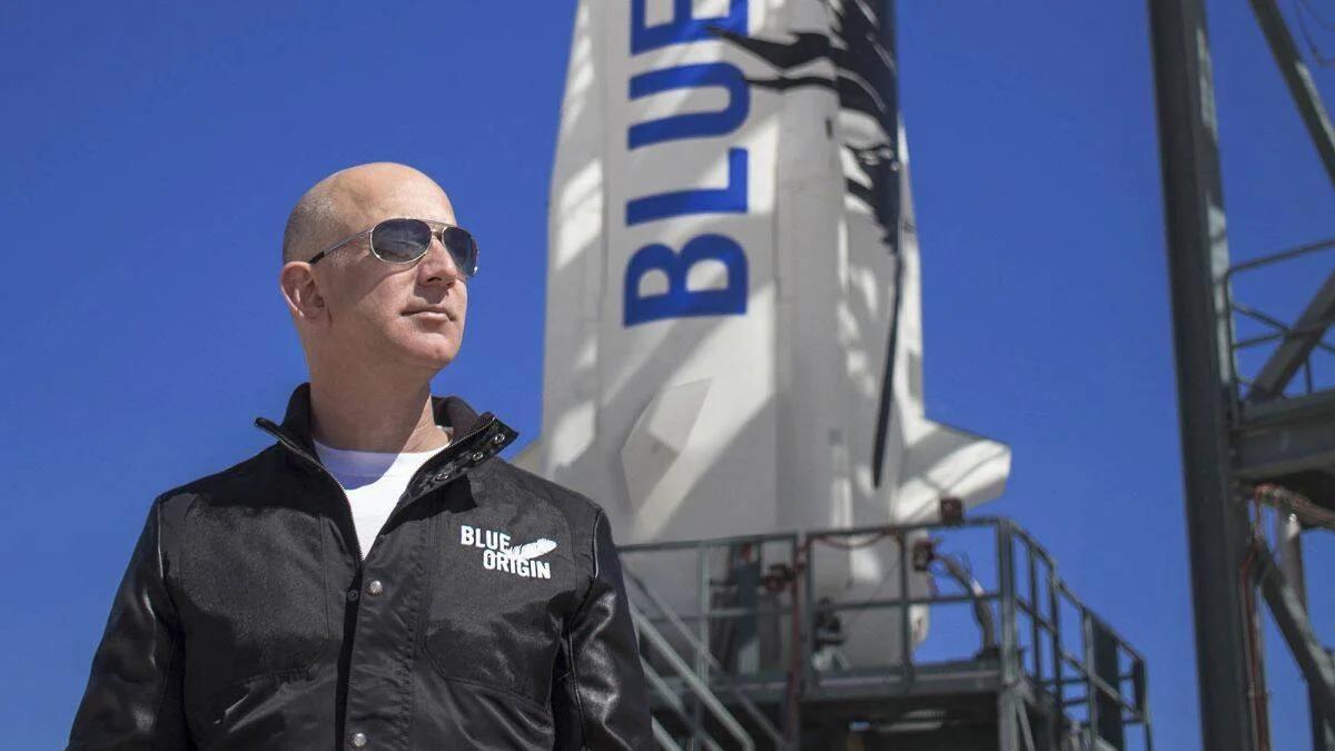 Hombre con gafas de sol y chaqueta de Blue Origin posando frente al cohete de la empresa espacial en un día soleado.