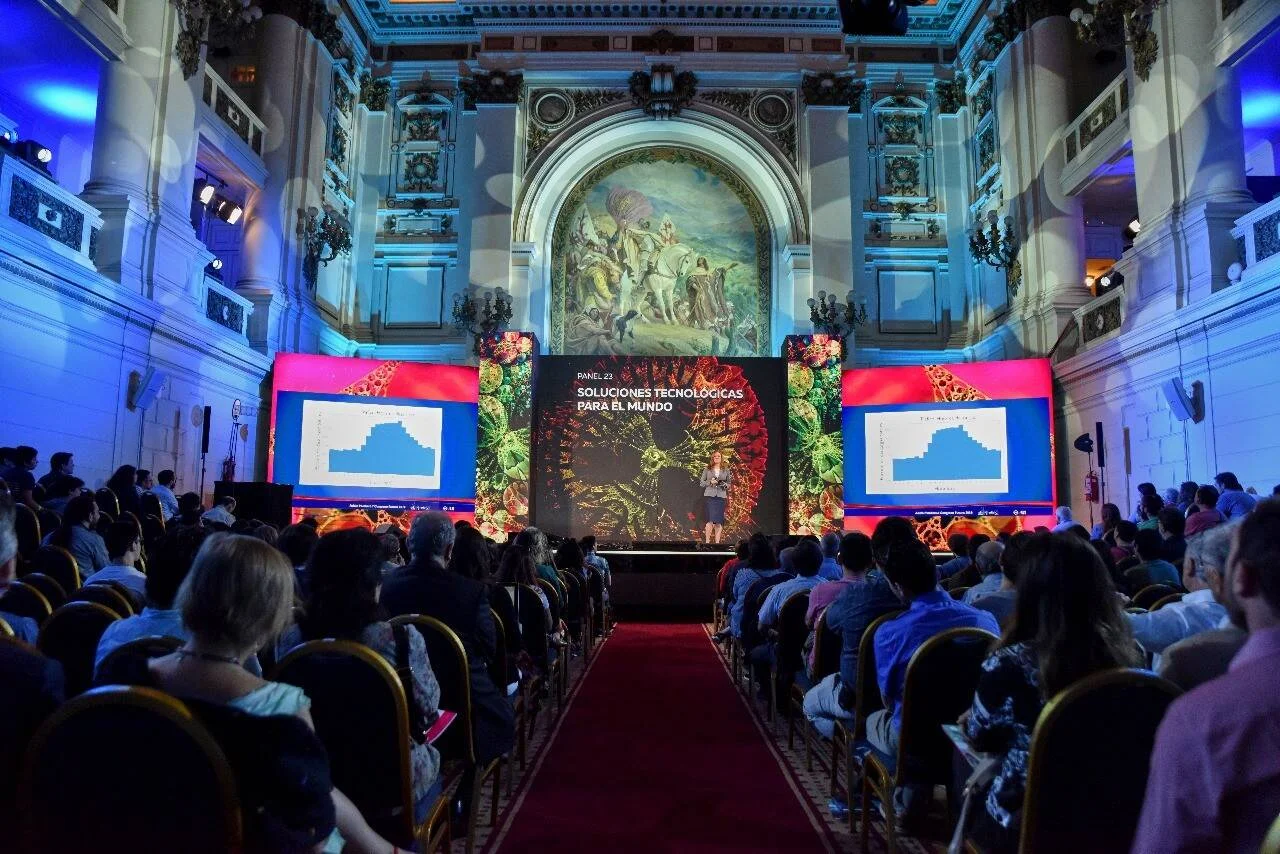 Oradora presenta el panel 'Soluciones Tecnológicas para el Mundo' ante un auditorio en un salón neoclásico con murales y pantallas LED.