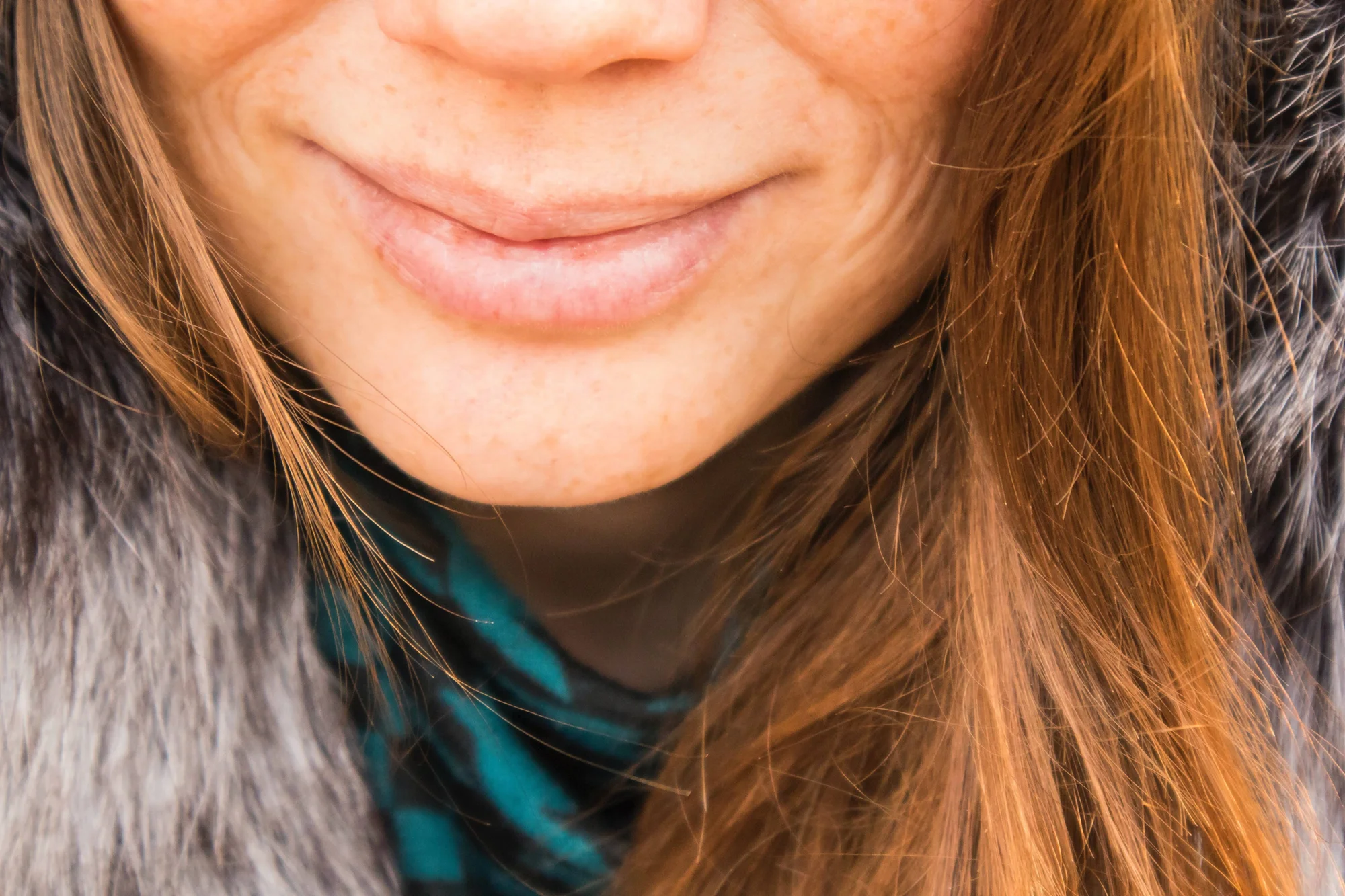 Primer plano de los labios y mentón de una mujer con cabello castaño, abrigo de piel gris y bufanda verde, esbozando una leve sonrisa.