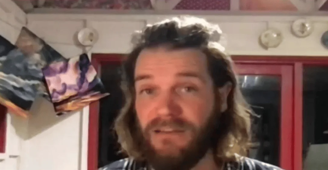 Hombre joven con cabello largo ondulado y barba habla frente a una cámara en un interior decorado con cuadros.