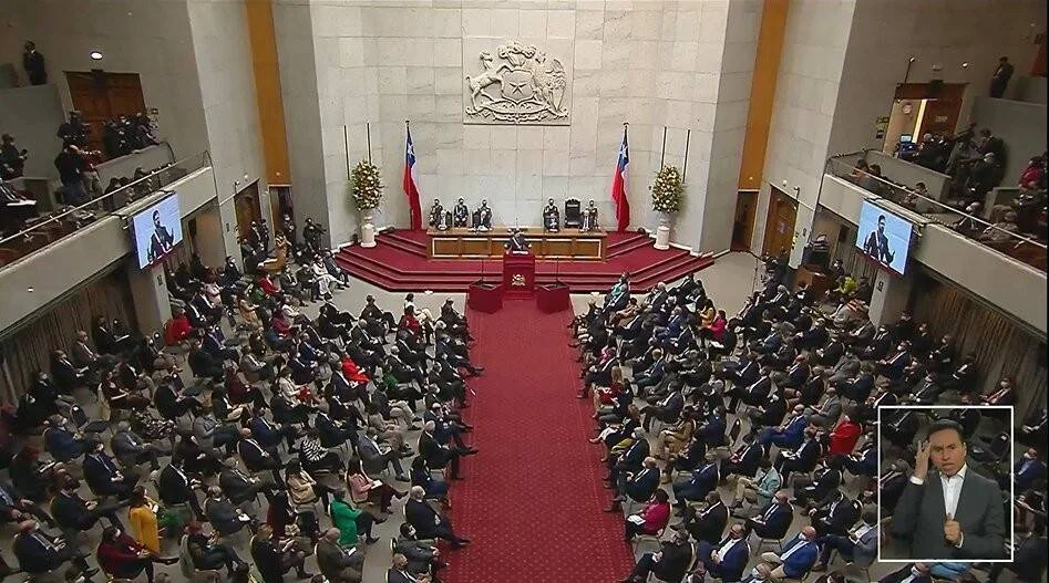Vista aérea del hemiciclo del Congreso de Chile repleto de autoridades, con un orador en el podio flanqueado por banderas chilenas y el escudo nacional al fondo.